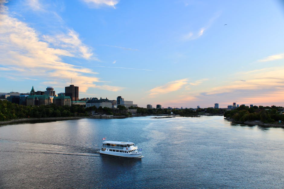 A touring boat cruises on a river with a city skyline backdrop under a bright sky.